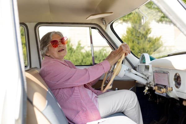 Elderly woman wearing pink sunglasses, driving a vintage car with a relaxed expression
