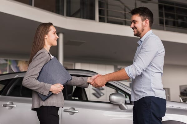 Customer shaking hands with a dealership representative next to a car, finalizing a discussing insurance option