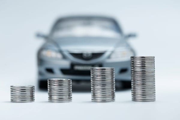 Stacks of coins increasing in size in front of a car, illustrating rising auto insurance costs due to inflation