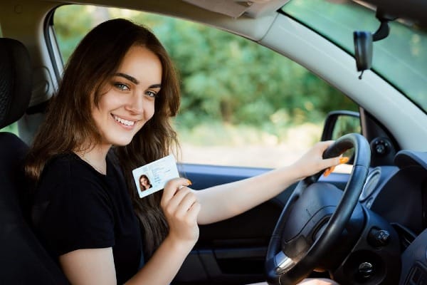 Young woman holding her driver’s license while sitting in the driver’s seat of a car, representing part-time driver auto insurance coverage